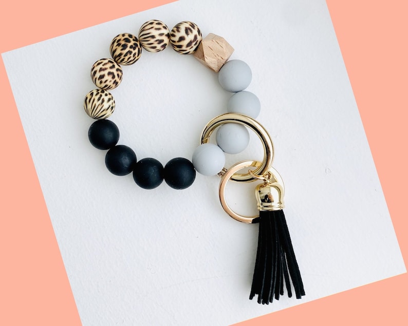 May include: A black, gray, and leopard print beaded keychain with a black tassel. The keychain is made of silicone beads and has a gold metal key ring.