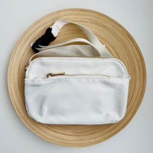 K&ouml;nnte beinhalten: Wei&szlig;e Bauchtasche mit goldenem Rei&szlig;verschluss und verstellbarem schwarzem Gurt. Die Bauchtasche liegt auf einer Holzoberfl&auml;che.