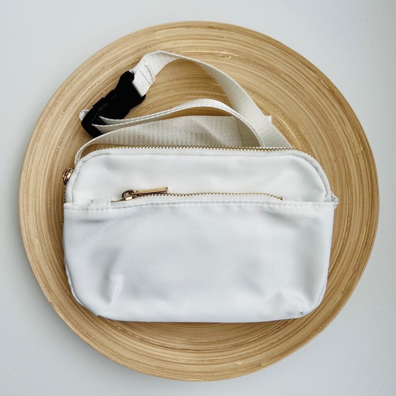 K&ouml;nnte beinhalten: Wei&szlig;e Bauchtasche mit goldenem Rei&szlig;verschluss und verstellbarem schwarzem Gurt. Die Bauchtasche liegt auf einer Holzoberfl&auml;che.
