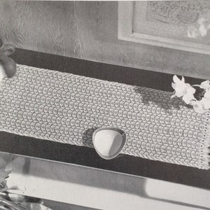 May include: A white crocheted table runner with a delicate lace pattern is displayed on a dark surface. A small white bowl and white flowers are also on the surface.
