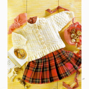 May include: A white knitted baby cardigan with a ruffled collar and button closure. The cardigan is paired with a red and black plaid skirt. The text "Patons PBN C 4863 To fit 0-3 Years 16-22 in DOUBLE KNITTING featured in FAIRYTALE DK" is printed on the image.
