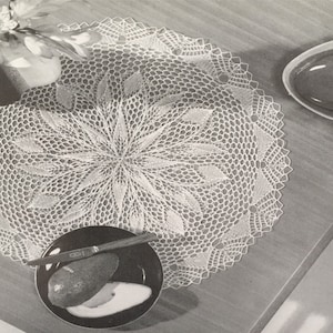 May include: A white crocheted doily with a delicate floral design. The doily is laid flat on a wooden table with a small bowl and a plate with a knife and fruit.