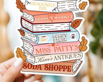 May include: A stack of colorful books with the names of fictional shops from the TV show "Gilmore Girls" printed on the spines. The books are arranged in a pile with the top book reading "Dragonfly Inn" and the bottom book reading "Soda Shoppe".
