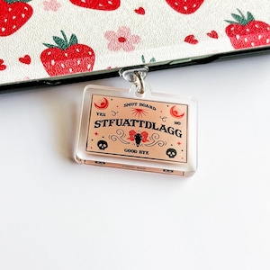 Puede incluir: Un amuleto de plástico rosa y blanco con el texto "SMUT BOARD" y "STFUATTDLAGG" en letras negras. El amuleto también presenta una mariposa negra, dos calaveras y una luna creciente.