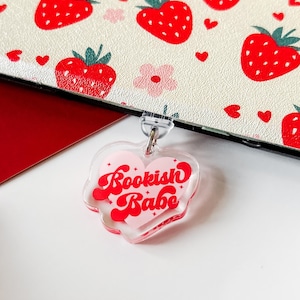 May include: A clear heart-shaped keychain with the text "Bookish Babe" in red script. The keychain hangs in front of a cream-colored surface with a strawberry pattern. The strawberries are red with green tops, and there are pink flowers and red hearts.