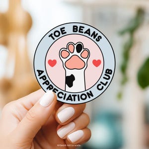 May include: A round sticker with the text "TOE BEANS APPRECIATION CLUB" encircling a pink circle with a cat paw print and two red hearts. The paw print has pink pads and black accents.