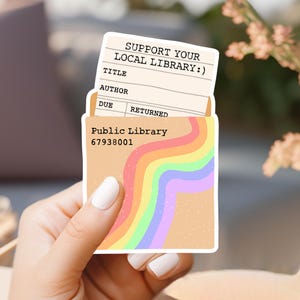 May include: A sticker with a rainbow design and the text "Support Your Local Library!" and "Public Library 67938001". The sticker is shaped like a library card with fields for "Title", "Author", "Due", and "Returned".