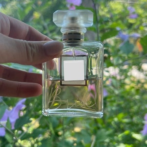 Peut inclure: Un flacon de parfum en verre transparent avec un bouchon doré et une étiquette blanche. Le flacon est posé sur une main, et l'arrière-plan est une image floue de feuilles vertes et de fleurs violettes.