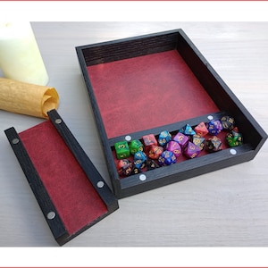 Puede incluir: Una bandeja de dados de madera negra con interior rojo, que contiene una variedad de dados poliédricos coloridos. La bandeja está abierta, con una tapa separada que también presenta un interior rojo. Un pergamino enrollado y una vela están en el fondo.