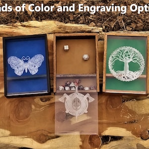 May include: Three wooden boxes with different coloured felt interiors and engraved designs. The boxes feature a butterfly, a tree of life, and a circle with axes. The boxes are on a wooden surface.