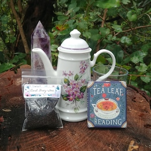 Puede incluir: Naturaleza muerta con una tetera blanca con detalles florales morados, una bolsa de té Earl Grey, una guía de lectura de hojas de té, un vaso y un cristal morado. Los objetos están dispuestos sobre una superficie de madera.