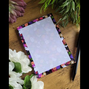 May include: A notepad with a black background and a white border. The border is decorated with a floral pattern in shades of purple, pink, orange, and yellow. The pattern includes ghosts and skulls. The notepad has a blank white page.