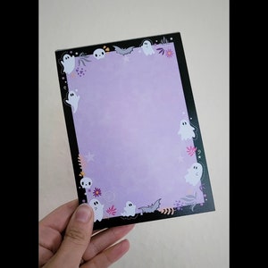May include: Purple notepad with a black border and a Halloween theme. The notepad has a ghost, bat, and flower design around the edges.