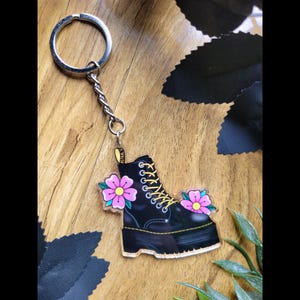 May include: A black boot-shaped keyring with yellow laces and pink flower details. The keyring has a silver ring and chain. The boot design features stitching and a platform sole. The background is a wooden surface.