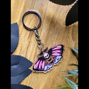 May include: A keychain with a moth design, featuring a skull on its head. The moth has pink and black wings with a crescent moon and dot pattern. The keychain is attached to a silver metal ring and chain.