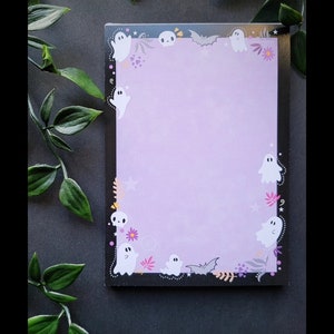 May include: A black and white notepad with a purple background and a border of ghosts, bats, and flowers. The notepad has a blank space in the center for writing.
