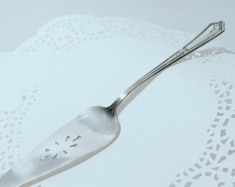 May include: A silver cake server with a decorative handle and a slotted blade. The blade has a starburst design. The server is resting on a white, lacy paper doily.