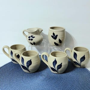 May include: Set of six stoneware mugs and a creamer, all with a cream base and blue leaf designs. The mugs have handles and the creamer has a spout.