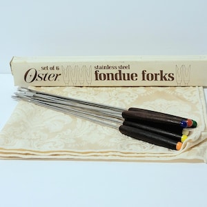 May include: A set of six stainless steel fondue forks with dark wooden handles and color-coded tips. The forks are displayed on a cream-colored patterned cloth, with the original box visible. The box reads "Oster" and "fondue forks."