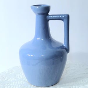 May include: A glossy periwinkle blue ceramic pitcher featuring a rounded base, a slender neck, a small spout, and a single angular handle attached to the neck and the upper body. The surface has a smooth, reflective finish with subtle light highlights.