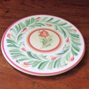 May include: A white ceramic plate with a hand-painted design of green leaves and a single orange flower. The plate has a red and yellow border.