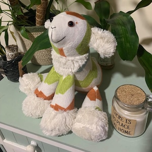 May include: A plush white, green, and orange chevron patterned lamb with a white fluffy face and brown eyes. The lamb is sitting on a light green surface with a candle jar in the background.
