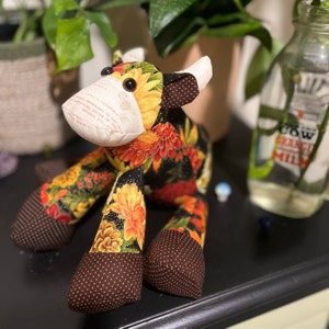May include: A plush cow toy with a black and white polka dot pattern on the ears and hooves. The body is made of a black fabric with a floral pattern in shades of red, yellow, and orange.