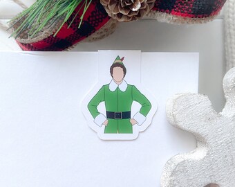 May include: A white sticker featuring a cartoon illustration of a character in a green elf costume with a white collar and cuffs, and a black belt. The sticker is placed on a white surface, with holiday decorations in the background.