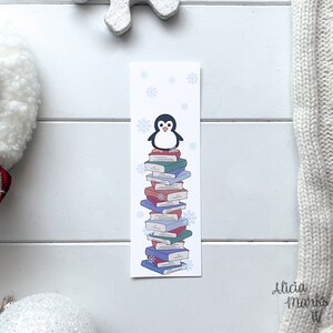 Peut inclure: Un marque-page blanc avec un pingouin debout sur une pile de livres colorés. Le pingouin est noir et blanc avec un bec jaune. L'arrière-plan est bleu clair avec des flocons de neige.