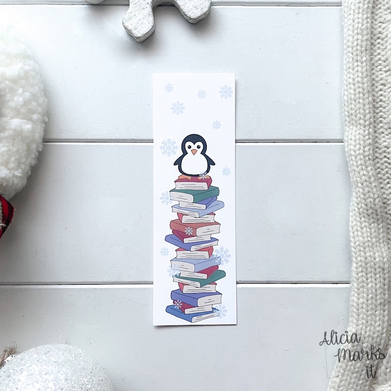 Pode incluir: Um marcador branco com um pinguim em p&eacute; em cima de uma pilha de livros coloridos. O pinguim &eacute; preto e branco com um bico amarelo. O fundo &eacute; azul claro com flocos de neve.