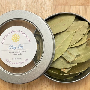 May include: An open, round, silver tin filled with dried bay leaves, next to the lid. The lid has a clear window with a label that reads "Girl Gone Herbal Botanicals Bay Leaf". The leaves are a light green color.