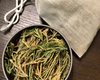 May include: A close-up of a small metal container filled with dried green and brown cedar leaves. The leaves are loose and ready to be used for crafting or aromatherapy.