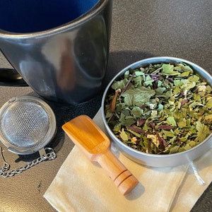 May include: A black mug with a blue interior sits next to a stainless steel tea infuser and a small wooden scoop. A metal tin holds dried tea leaves, and a small cloth bag is also present. The tea leaves are a mix of green and purple.