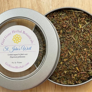 May include: A silver tin of dried St. John's Wort, with the lid partially open. The label reads "Girl Gone Herbal Botanicals" and "St. John's Wort". The tin contains 113 grams by volume of the certified organic herb.