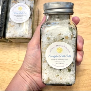 Puede incluir: Un frasco de vidrio lleno de sales de baño blancas y hierbas verdes. El frasco tiene una tapa plateada y una etiqueta blanca que dice "Girl Gone Herbal Botanicals Eucalyptus Bath Salts Epsom salt, eucalyptus leaves, organic sweet mint GirlGoneHerbal.com"