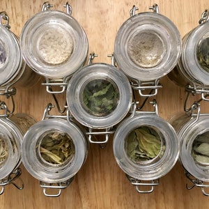 May include: Eight clear glass jars with metal lids, filled with various dried herbs and spices, are arranged in two rows of four on a wooden surface.
