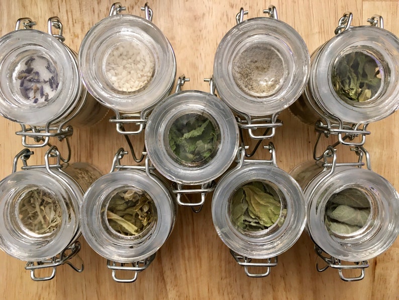 May include: Eight clear glass jars with metal lids, filled with various dried herbs and spices, are arranged in two rows of four on a wooden surface.