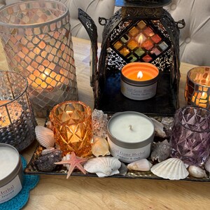May include: A collection of lit candles in decorative holders and tins, arranged on a tray with seashells and stones. The candles are in various colors, including orange, purple, and clear glass. One candle tin reads "Girl Gone Herbal".