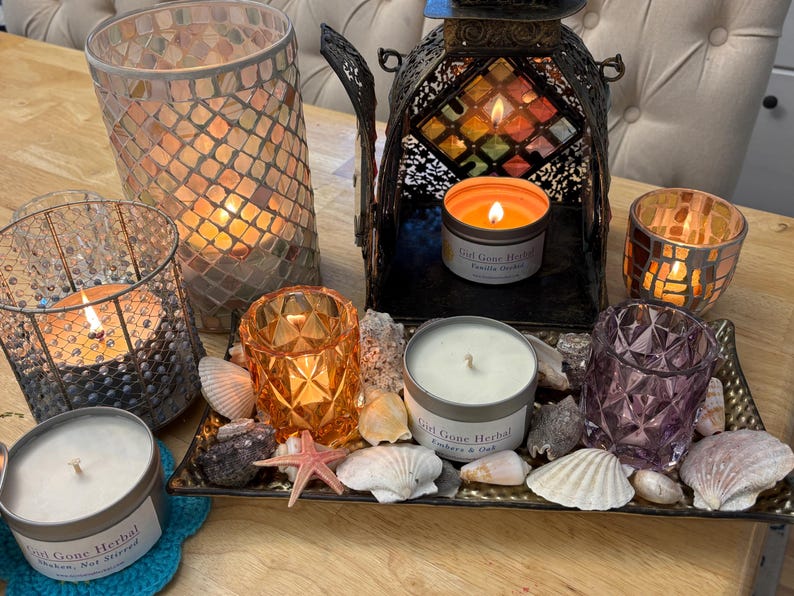 May include: A collection of lit candles in decorative holders and tins, arranged on a tray with seashells and stones. The candles are in various colors, including orange, purple, and clear glass. One candle tin reads "Girl Gone Herbal".