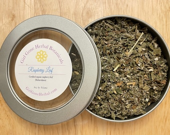 Op de afbeelding: Een zilveren blik met een doorzichtig deksel gevuld met gedroogde frambozenbladeren. Het etiket op het blik luidt "Girl Gone Herbal Botanicals Raspberry Leaf Certified organic raspberry leaf (Rubus idaeus) 4oz by Volume GirlGoneHerbal.com"