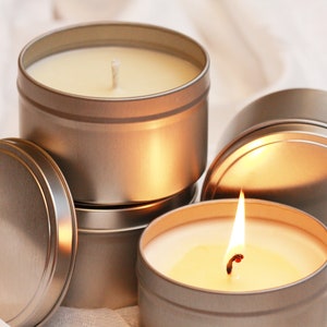 May include: Several silver-colored, round tin candles, some with lids. One candle is lit, showing a bright flame. The candles are filled with a creamy wax and have white wicks. The background is a soft, white fabric.