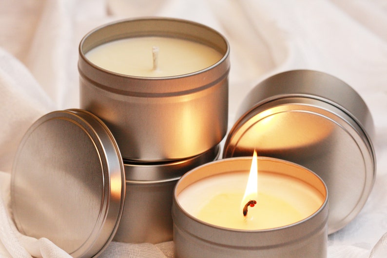 May include: Several silver-colored, round tin candles, some with lids. One candle is lit, showing a bright flame. The candles are filled with a creamy wax and have white wicks. The background is a soft, white fabric.