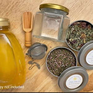 May include: A set of herbal tea blends in silver tins with clear lids. The tins are labeled "Gift Gone Herbal Remedies" and "Heather Herbal Tea". There is also a glass jar with a gold lid, a wooden scoop, a metal tea infuser, and a bottle of honey. Honey not included.