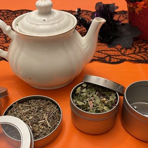 May include: A white teapot with a gold trim sits on an orange surface with three silver tins. One tin is open and contains dried herbs. Another tin is open and contains dark berries. The third tin is closed.