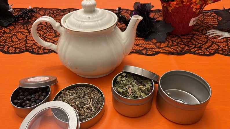 May include: A white teapot with a gold trim sits on an orange surface with three silver tins. One tin is open and contains dried herbs. Another tin is open and contains dark berries. The third tin is closed.