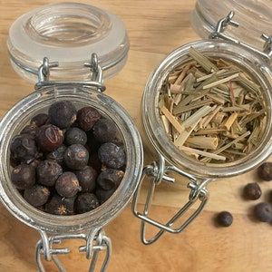 May include: Two glass jars with metal lids, one filled with dried lemongrass and the other with dark berries. The jars are on a wooden surface with a few berries scattered around.