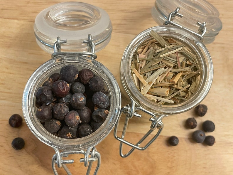 May include: Two glass jars with metal lids, one filled with dried lemongrass and the other with dark berries. The jars are on a wooden surface with a few berries scattered around.