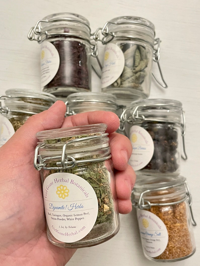 May include: Six small glass jars with metal lids, filled with various herbs and spices. The jars are labelled with the text "Gid Gone Herbal Botanicals" and "Dynamite! Herbs". The label also lists the ingredients: "Bul Tarragon, Organic Lemon Peel, Onion Powder, White Pepper".