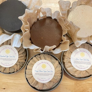 Puede incluir: Cinco barras redondas de exfoliante de azúcar en latas de metal con envoltorios de papel marrón. Las barras están etiquetadas con el texto "Girl Gone Herbal Botanicals" y los nombres de las fragancias: "Ground Coffee", "Orange Blossom", "Lemon Sugar", "Grapefruit" y "Vanilla Bean".