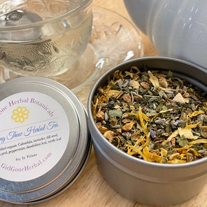 May include: A silver tin with a white label that reads "Girl Gone Herbal Botanicals Spring Thaw Herbal Tea" and lists the ingredients. The tin is filled with a blend of dried herbs, including lavender, calendula, lemon peel, and peppermint.